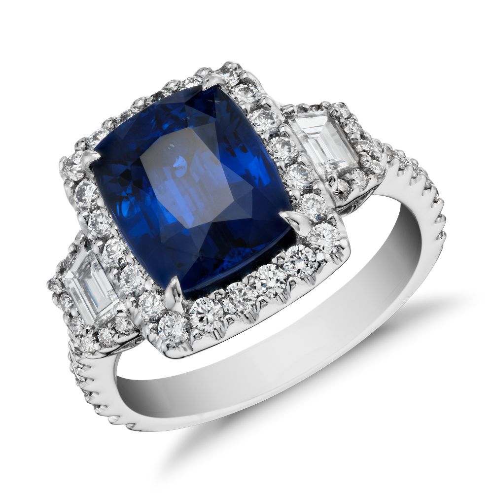 Sapphire and Diamond Halo Three-Stone Ring in 18k White Gold (3.65 ct) (9.8x7.8mm) Distinctly glamorous, this sapphire gemstone ring showcases a 3.65 ct cushion sapphire, step cut trapezoid side stones framed by a halo of brilliant pavé diamonds and a filigree flourish set in enduring 18k white gold. 