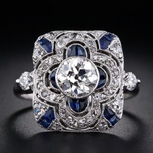 This is an exquisite dinner ring from the early 20th century. This early Art Deco ring centers an .80 carat European cut diamond bezel set in a rectangular plaque which is delicately pierced and set with both glittering antique cut diamonds as well as calibre cut synthetic sapphires. A round cut diamond highlights each shoulder.